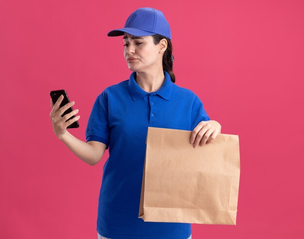 Mujer de entrega joven disgustada en uniforme y gorra sosteniendo el paquete de papel y el teléfono móvil mirando el teléfono aislado en la pared rosa