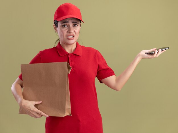 Mujer de entrega joven irritada en uniforme y gorra con paquete de papel y teléfono móvil aislado en la pared verde oliva