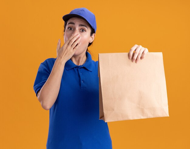 Mujer de entrega joven preocupada en uniforme y gorra sosteniendo el paquete de papel manteniendo la mano en la boca