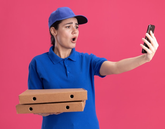 Mujer de entrega joven preocupada en uniforme y gorra sosteniendo paquetes de pizza tomando selfie