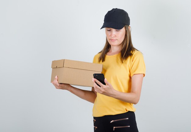 Mujer de entrega mirando smartphone con caja de cartón en camiseta, pantalón y gorra.