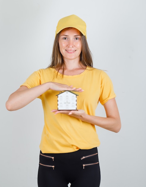 Foto gratuita mujer de entrega con modelo de casa en camiseta, pantalón, gorra y aspecto alegre.