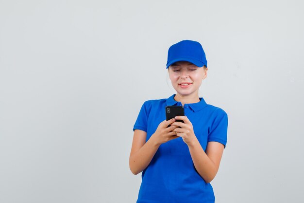 Mujer de entrega que tiene chat de texto en el teléfono móvil en camiseta azul y gorra y mirando alegre