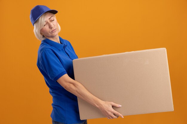 Mujer de entrega rubia de mediana edad cansada en uniforme azul y gorra de pie en la vista de perfil sosteniendo cardbox con los ojos cerrados aislados en la pared naranja