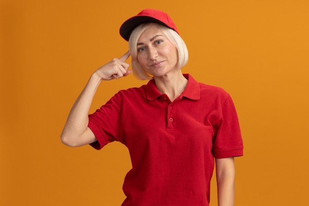 Mujer de entrega rubia de mediana edad complacida en uniforme rojo y gorra haciendo gesto de pensar aislado en la pared naranja