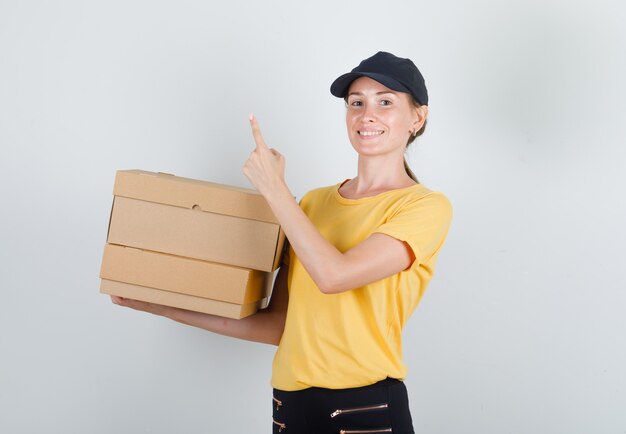 Mujer de entrega sosteniendo cajas de cartón con el dedo hacia arriba en camiseta, pantalón y gorra y mirando alegre