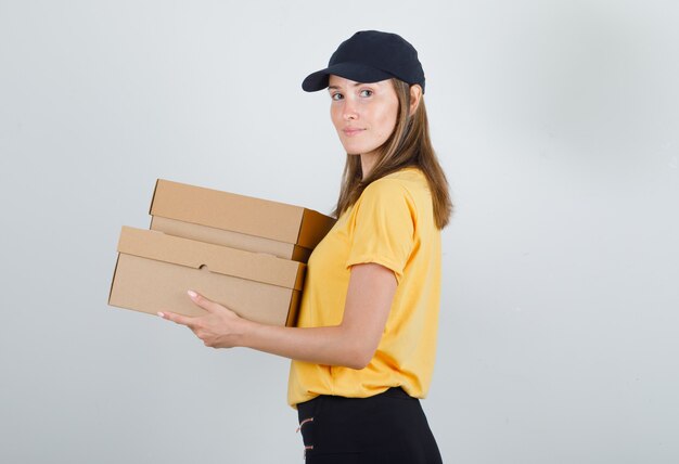 Mujer de entrega sosteniendo cajas de cartón y sonriendo en camiseta, pantalón y gorra.