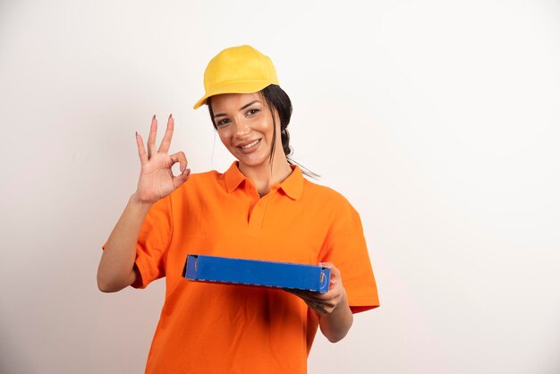 Mujer de entrega sosteniendo pizza y mostrando gesto ok en la pared blanca.