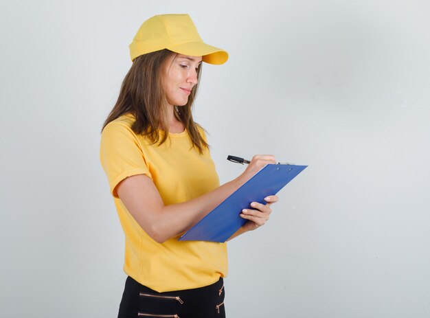 Mujer de entrega tomando notas en el portapapeles en camiseta, pantalón, gorra y mirando ocupado