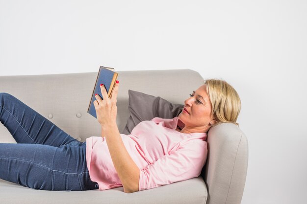 Mujer envejecida en blusa rosa leyendo libro en sofá