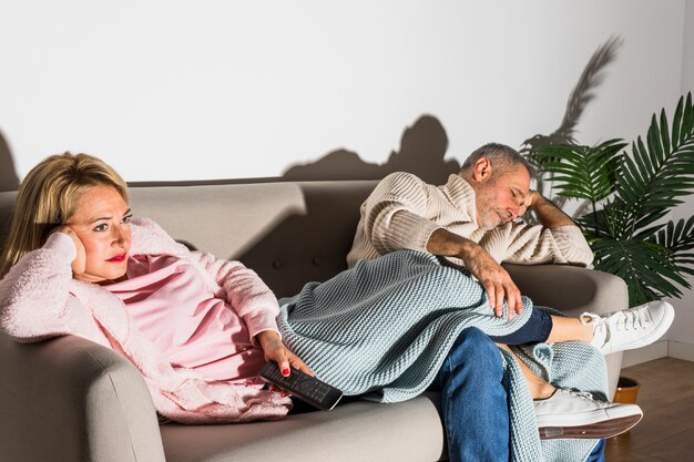 Mujer envejecida con control remoto de TV y hombre viendo TV en el sofá.