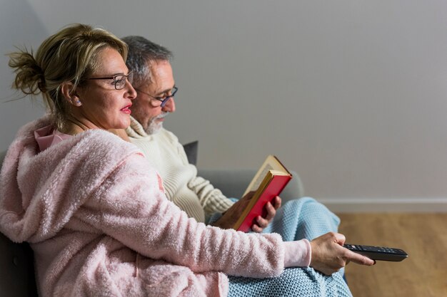 Mujer envejecida con control remoto de TV viendo televisión y libro de lectura del hombre en el sofá