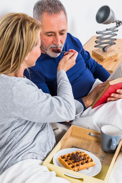 Foto gratuita mujer envejecida que da bayas al hombre con el libro cerca de la bandeja con desayuno en la cama