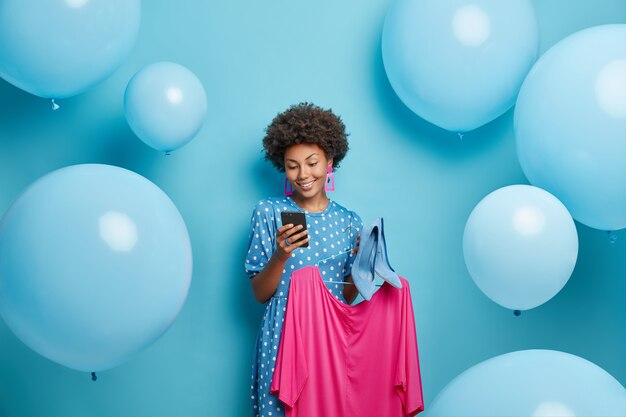 Mujer envía mensajes de texto a través de un teléfono inteligente tiene vestido rosa en la percha y zapatos de tacón se prepara para una ocasión especial aislado en azul