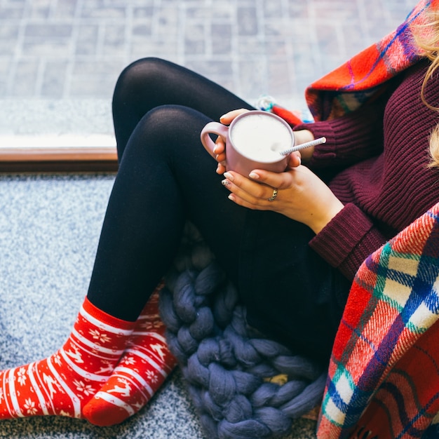 Mujer envuelta en tela escocesa se sienta en el suelo con una taza de chocolate caliente