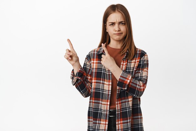 Mujer escéptica frunce el ceño, sonríe y entrecierra los ojos molestos, apuntando a la esquina superior izquierda molesta, no le gusta algo malo, no está segura, expresa su incredulidad, pared blanca