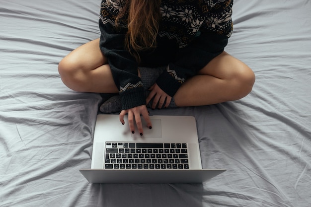 Mujer escribiendo en la computadora portátil mientras está sentado en la cama con las piernas cruzadas