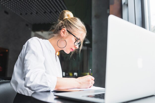 Mujer escribiendo en el documento con la computadora portátil en el escritorio