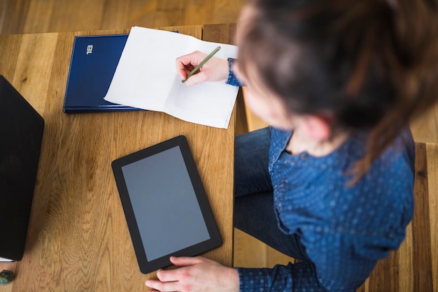 Mujer escribiendo notas sobre papel con tableta digital sobre escritorio de madera