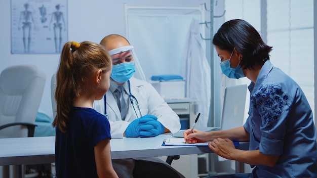 Mujer escribiendo prescripción en el portapapeles escuchando instrucciones del médico. Pediatra especialista en medicina con máscara que brinda servicios de atención médica, consulta, tratamiento en el hospital durante el covid-19