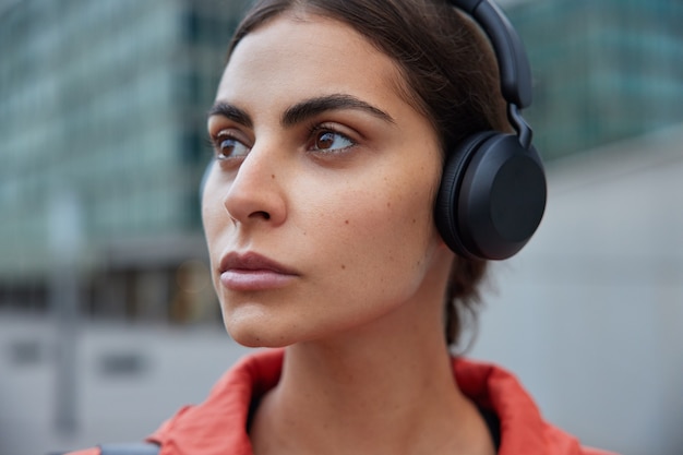 Mujer escucha la pista de audio en auriculares inalámbricos soñando despierto mientras camina al aire libre piensa en nuevos logros deportivos plantea contra borrosa