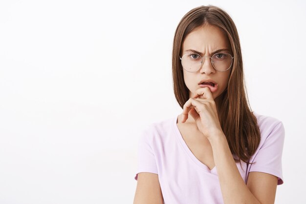 Mujer escuchando una extraña historia sospechosa, perpleja y dudosa, frunciendo el ceño, tocando el labio y mirando con incredulidad y mirada preocupada de pie con gafas y blusa sobre una pared blanca