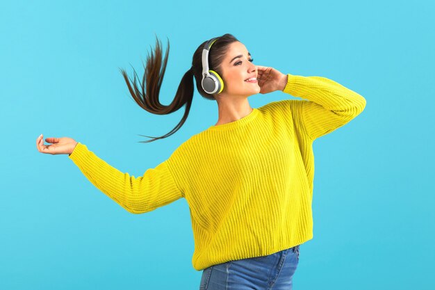 Mujer escuchando música en auriculares inalámbricos feliz vistiendo suéter tejido amarillo posando sobre azul