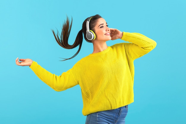 Mujer escuchando música en auriculares inalámbricos feliz vistiendo suéter tejido amarillo posando sobre azul
