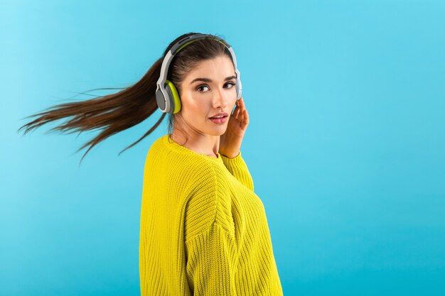 Mujer escuchando música en auriculares inalámbricos feliz vistiendo suéter tejido amarillo posando sobre azul