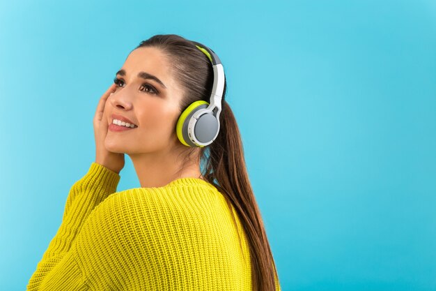 Mujer escuchando música en auriculares inalámbricos feliz vistiendo suéter tejido amarillo posando sobre azul