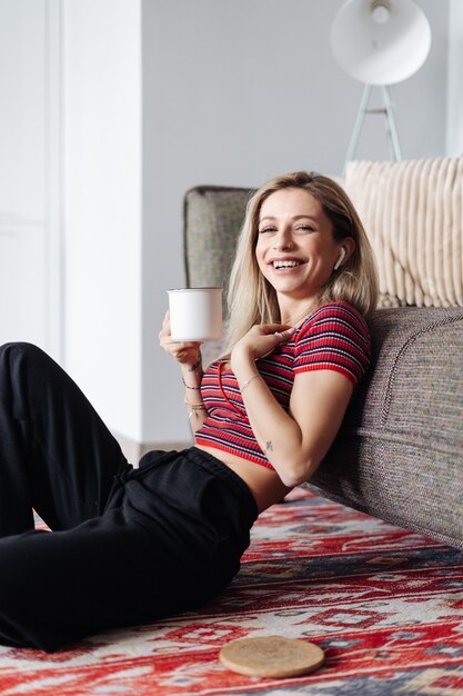 Mujer escuchando música en auriculares mientras está sentado cerca del sofá en la sala de estar bebiendo té