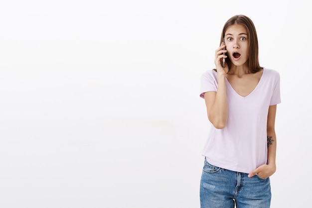 Mujer escuchando los últimos rumores frescos y destacados hablando por teléfono, sorprendida y sorprendida abriendo la boca de asombro y sorpresa sosteniendo el teléfono inteligente cerca de la oreja