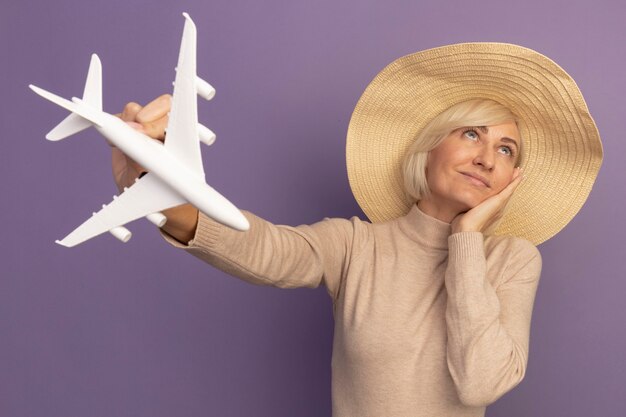 Foto gratuita mujer eslava bastante rubia complacida con sombrero de playa pone la mano en la cara sostiene el modelo de avión mirando hacia arriba en púrpura