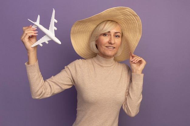 Mujer eslava bastante rubia complacida con sombrero de playa sosteniendo avión modelo en púrpura