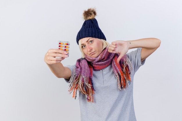 Mujer eslava enferma rubia joven disgustada con gorro de invierno y bufanda con los pulgares hacia abajo y sostiene el paquete de píldoras médicas aisladas en la pared blanca con espacio de copia