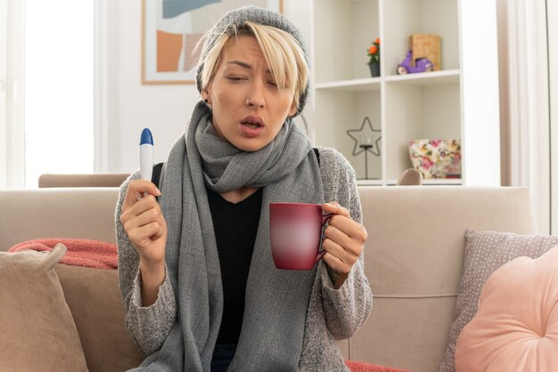 Mujer eslava joven enferma con bufanda alrededor de su cuello con sombrero de invierno sosteniendo termómetro y taza sentado en el sofá en la sala de estar