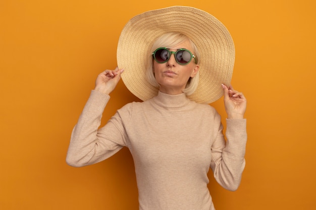 Mujer eslava rubia bastante segura con sombrero de playa en gafas de sol se encuentra en naranja
