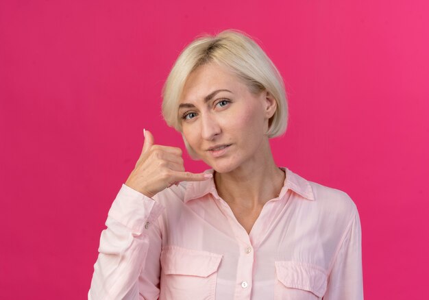 Mujer eslava rubia joven complacida mirando a cámara y haciendo gesto de llamada aislado sobre fondo rosa