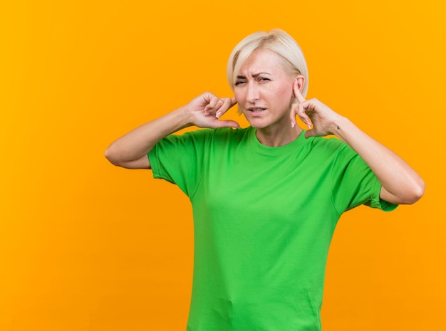 Mujer eslava rubia de mediana edad con el ceño fruncido mirando a la cámara poniendo los dedos en las orejas aisladas sobre fondo amarillo con espacio de copia