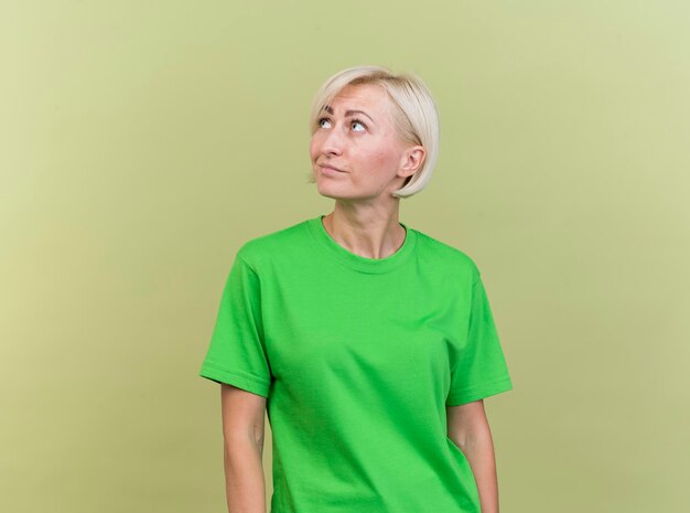 Mujer eslava rubia de mediana edad complacida girando de cabeza a lado mirando hacia arriba aislado en la pared verde oliva con espacio de copia