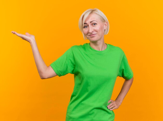 Mujer eslava rubia de mediana edad complacida mirando al frente manteniendo la mano en la cintura apuntando con la otra mano al lado aislado en la pared amarilla