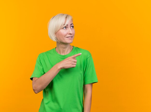 Mujer eslava rubia de mediana edad complacida mirando y apuntando al lado aislado sobre fondo amarillo con espacio de copia