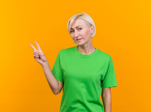 Mujer eslava rubia de mediana edad complacida mirando a la cámara haciendo el signo de la paz aislado sobre fondo amarillo con espacio de copia