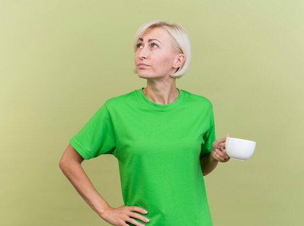 Mujer eslava rubia de mediana edad pensativa sosteniendo una taza de té manteniendo la mano en la cintura mirando al lado aislado en la pared verde oliva