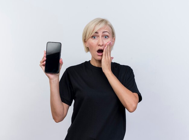 Mujer eslava rubia de mediana edad sorprendida mostrando teléfono móvil manteniendo la mano en la mejilla mirando a cámara aislada sobre fondo blanco con espacio de copia