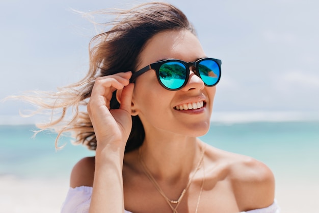 Mujer espectacular en gafas de sol brillantes de moda disfrutando de un buen día en el resort oceánico. Retrato al aire libre de mujer bronceada posando en la costa del mar en la mañana de verano.