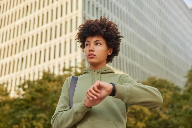 mujer espera al entrenador al aire libre verifica el tiempo en el reloj de pulsera vestida con poses de sudadera contra el edificio de la ciudad moderna en el exterior hace ejercicio con regularidad. Estilo de vida saludable