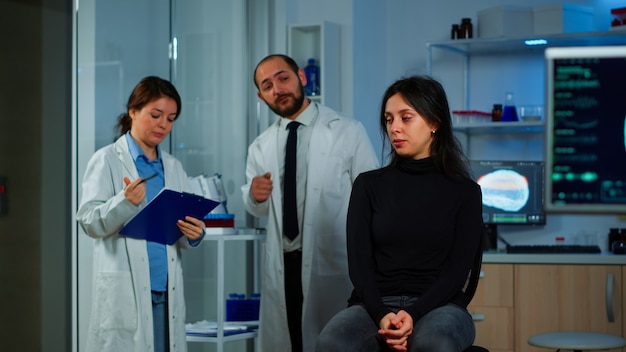 Mujer esperando médico sentado en una silla en el laboratorio de investigación neurológica, mientras que el equipo de investigadores discute en segundo plano el estado de salud del paciente, funciones cerebrales, sistema nervioso, tomografía