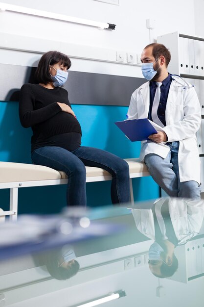 Mujer esperando a un niño reuniéndose con un especialista en salud en el consultorio, recibiendo consulta en una cita médica durante la pandemia de covid 19. Paciente expectante y médico discutiendo sobre el embarazo.