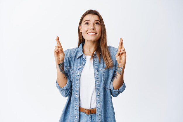 Mujer esperanzada cruza los dedos para la buena suerte y mirando hacia arriba con cara soñadora, pidiendo un deseo, de pie emocionado contra la pared blanca